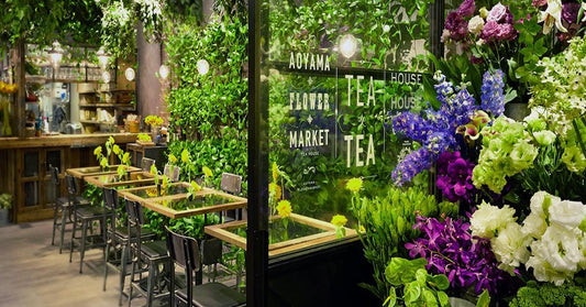 A cozy tea house filled with vibrant flower arrangements and lush green plants, featuring wooden tables with glass tops and warm ambient lighting — creating a peaceful, nature-inspired café atmosphere.